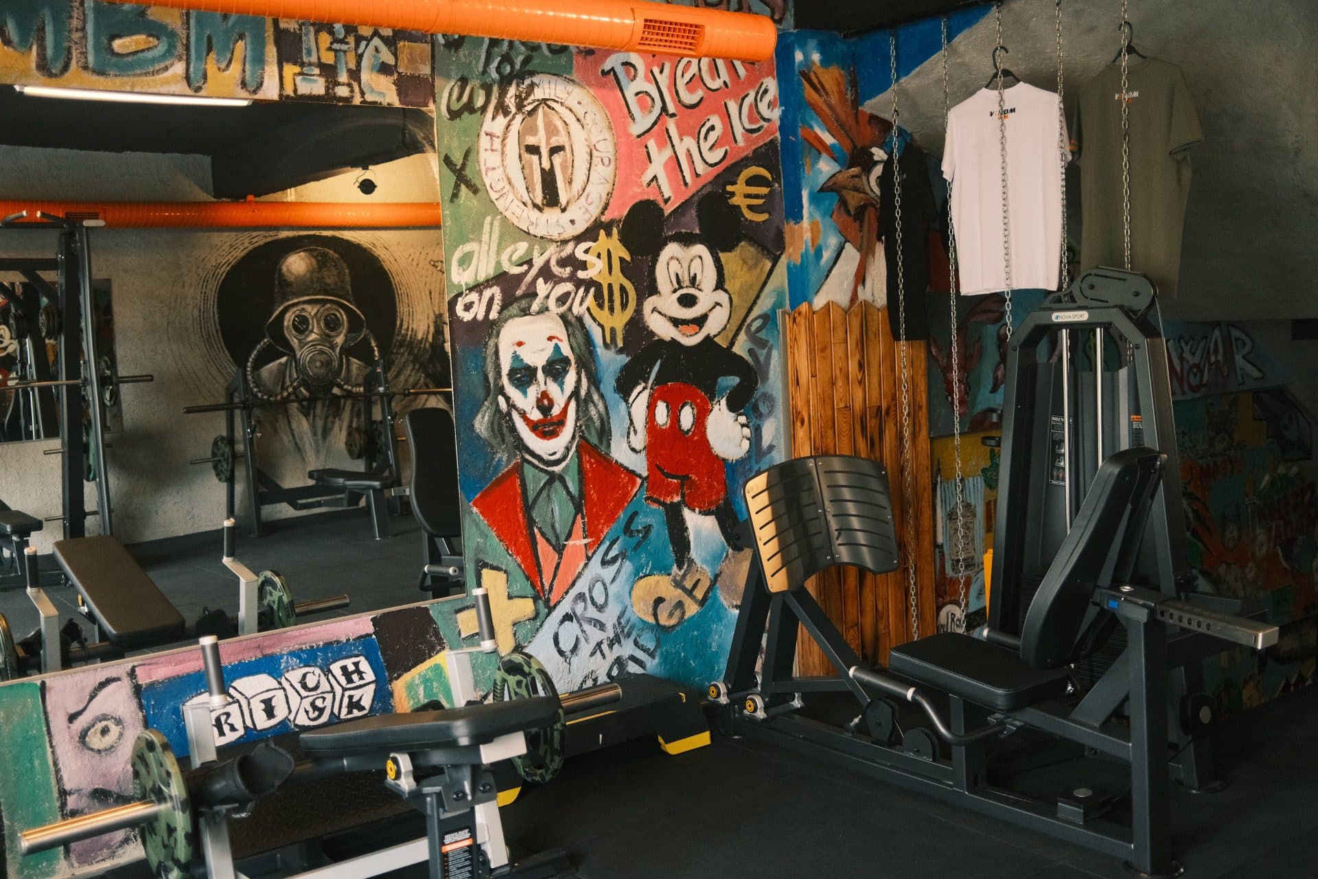 Joker portresi, Mickey Mouse ve Spartan miğferi graffitileriyle kaplı renkli duvar; önünde bacak press sledi ve asılı Venom Gym tişörtleri